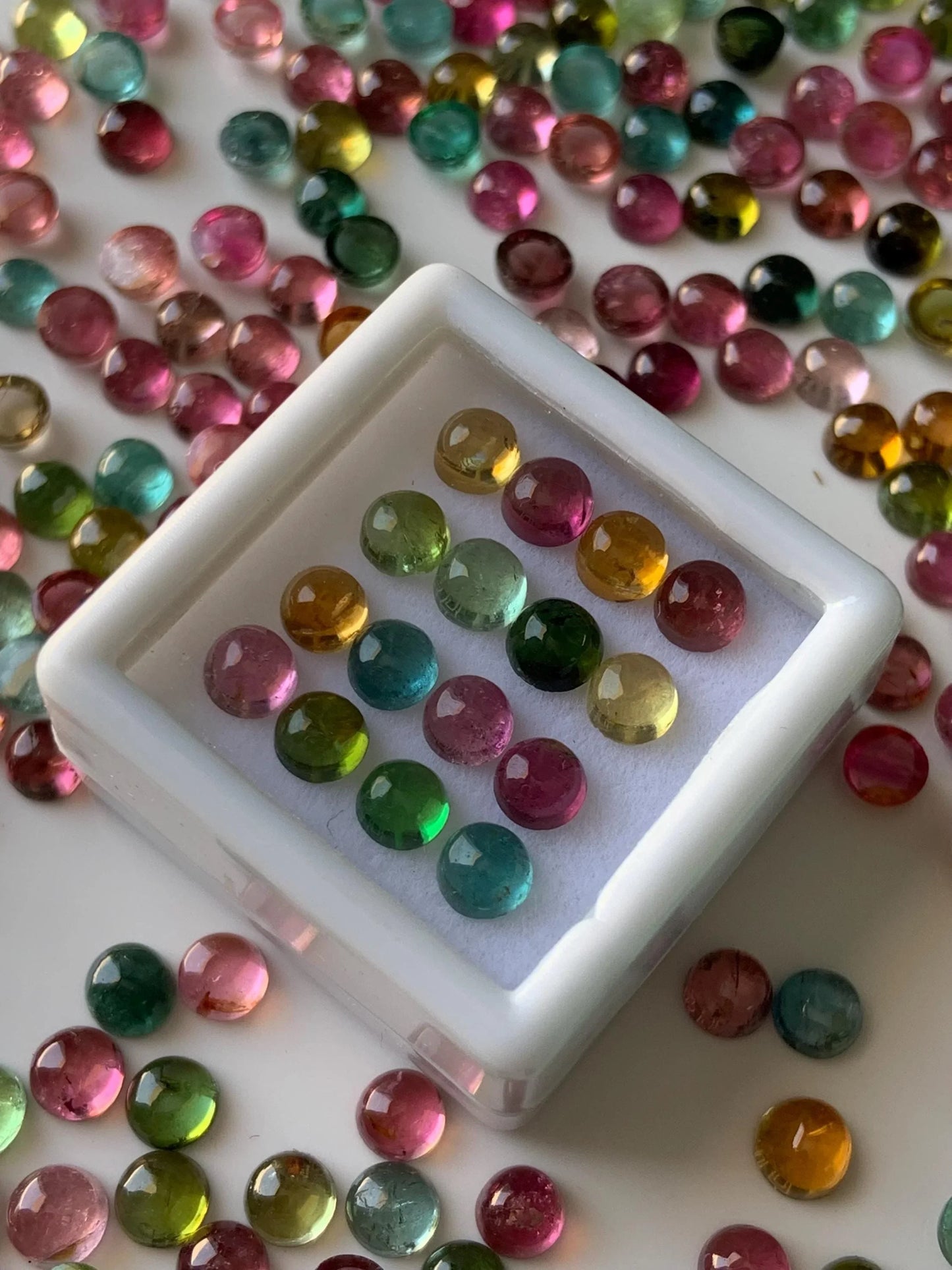 Multicolor round tourmaline cabochon gemstones arranged in display box and scattered on white surface