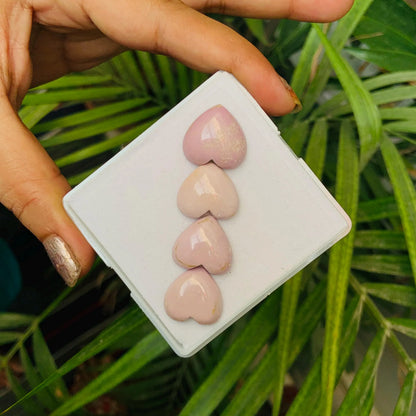 Hand holding four polished pink heart gemstone cabochons on white box, greenery background
