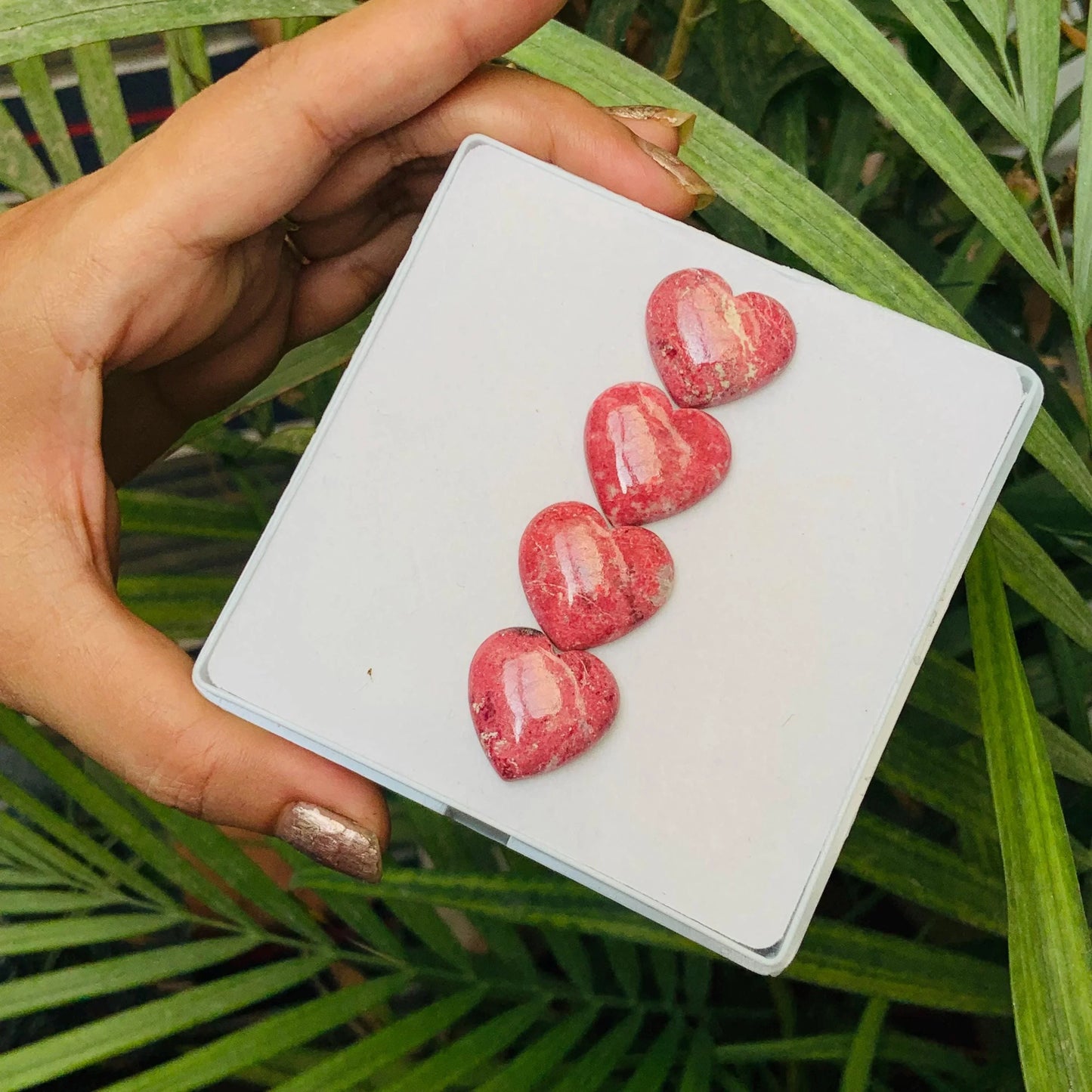 Hand holding display tray with four pink heart-shaped gemstone cabochons, outdoor background.