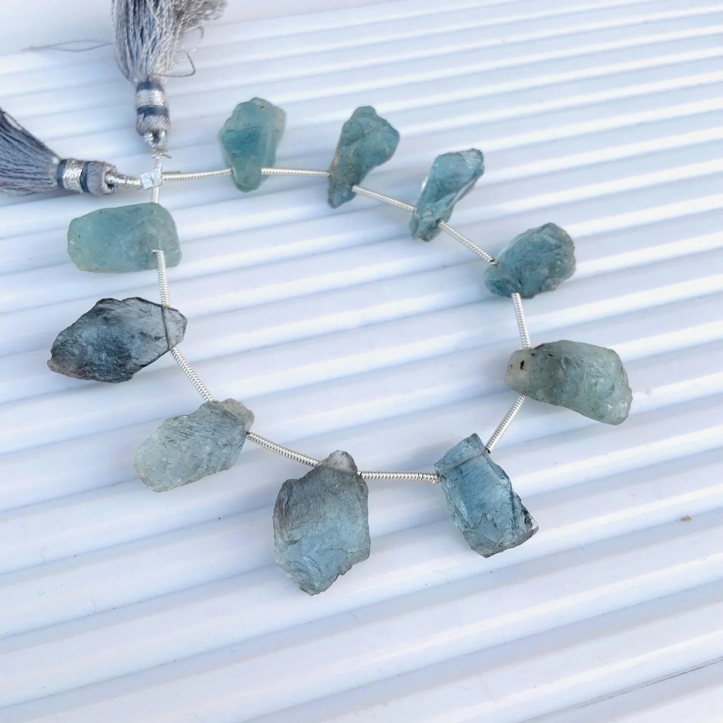 Strand of raw aquamarine gemstone beads with grey silk tassels on a white background