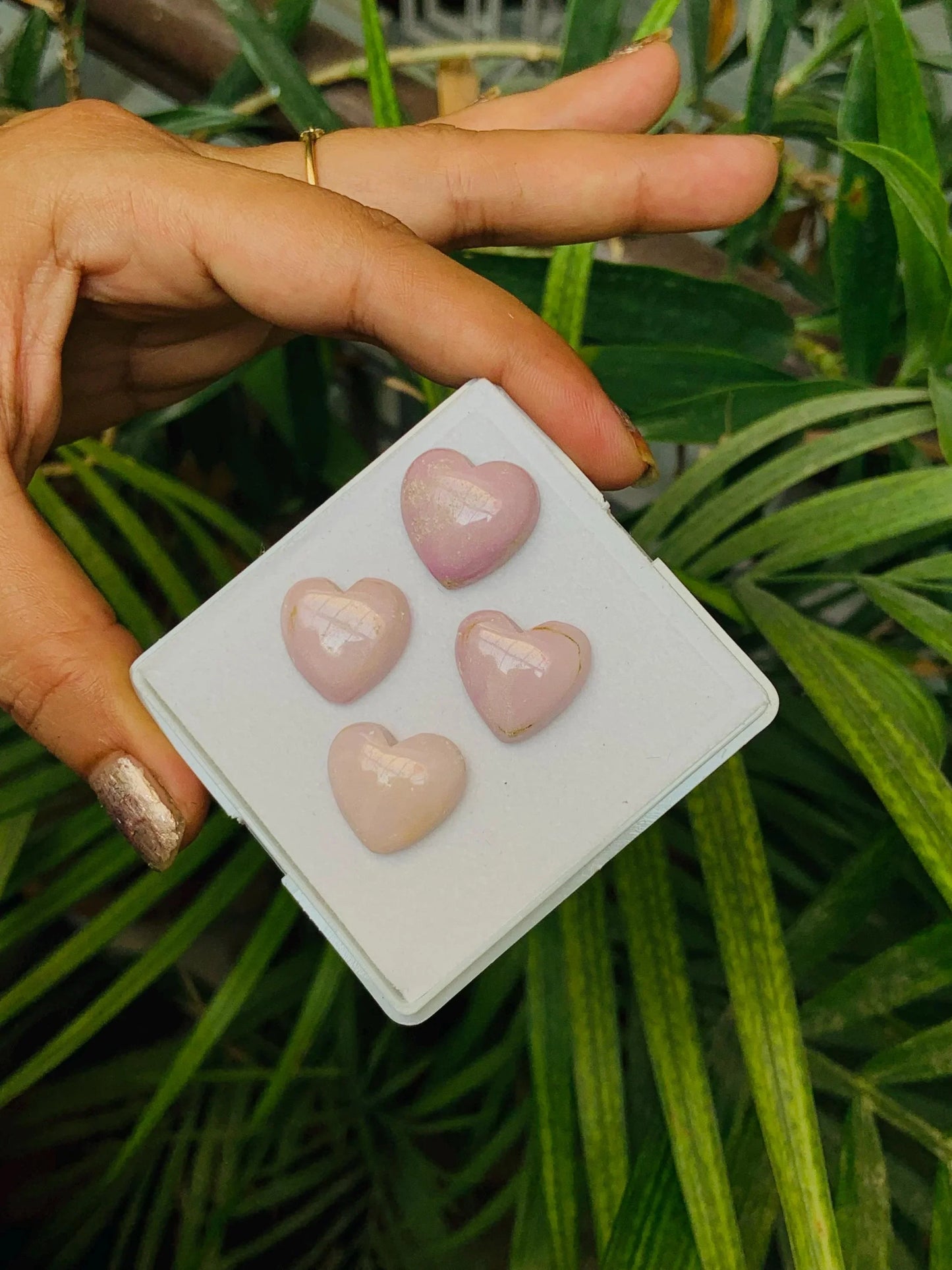 Hand holding tray of four heart-shaped pink gemstone cabochons with plants in background