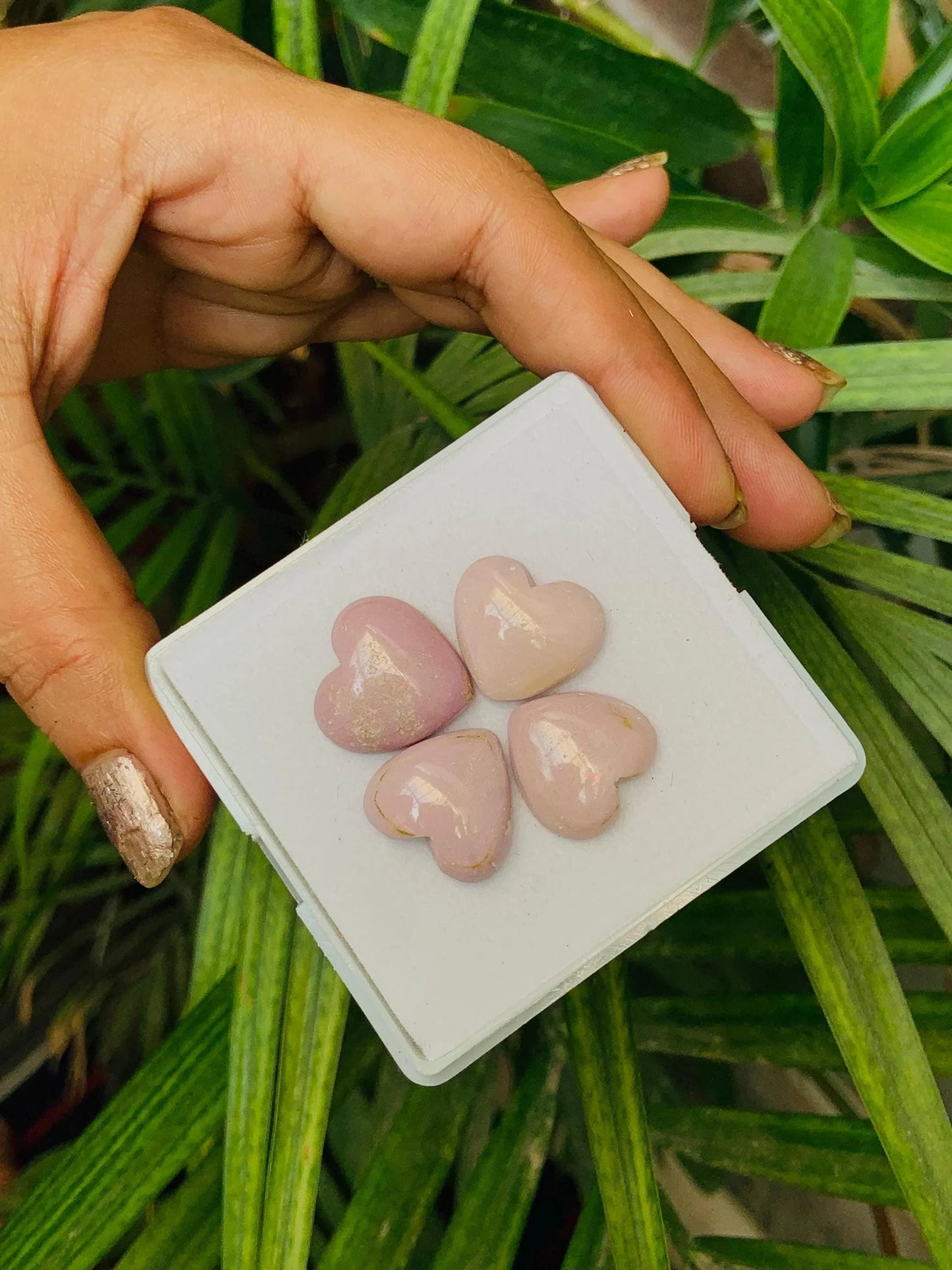 Hand holding four pink heart-shaped gemstone cabochons in display box, green leaves background