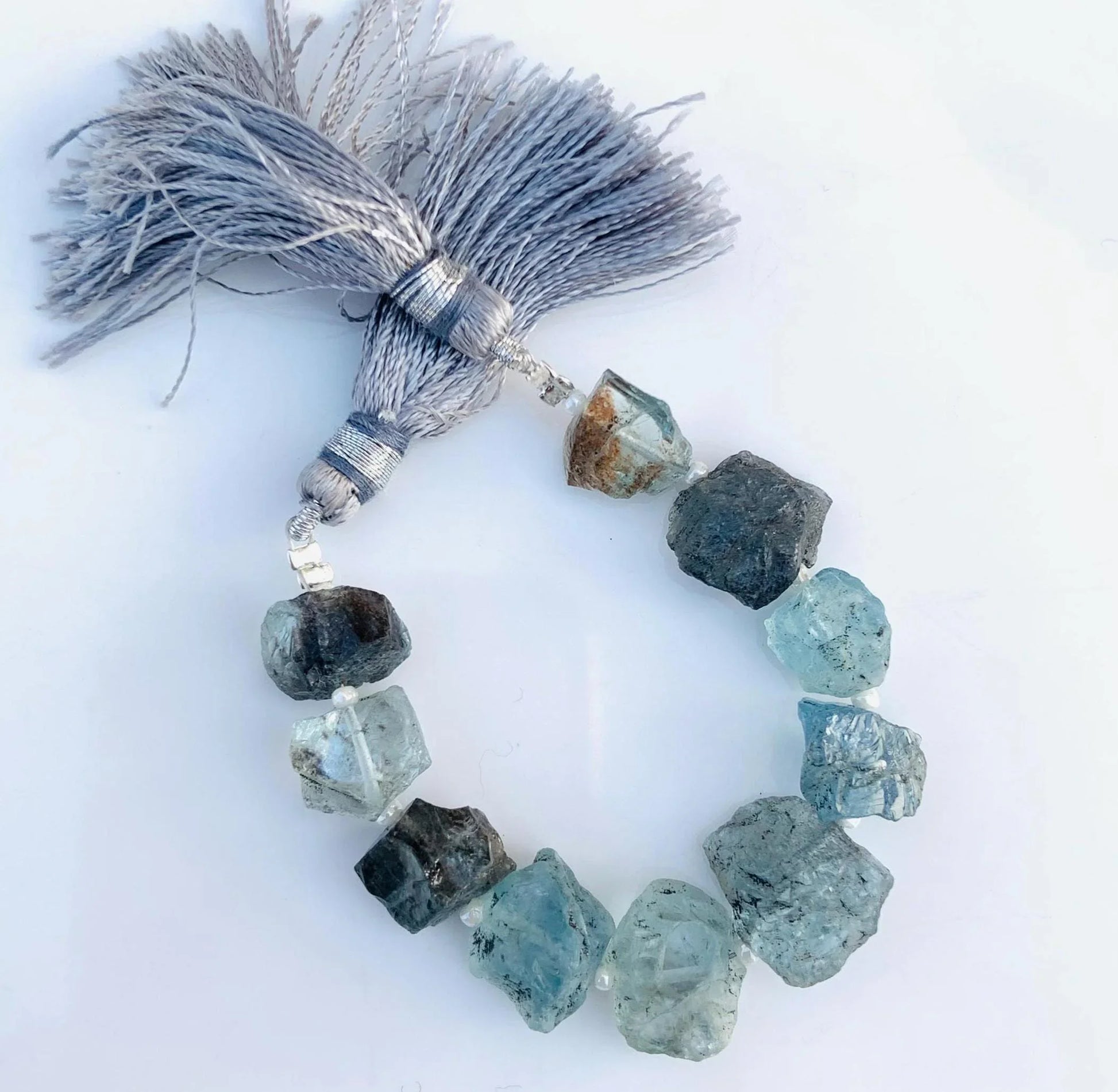 Raw aquamarine gemstone beads strand with grey tassels on white background