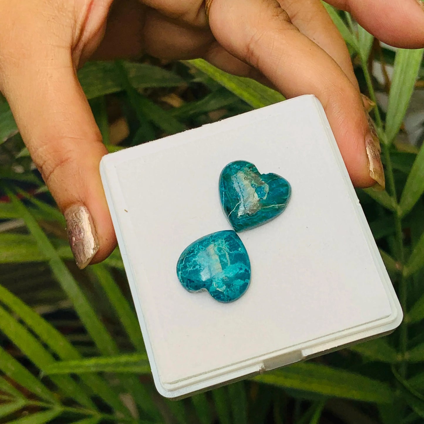 Hand holding two heart-shaped blue-green gemstone cabochons on a white box