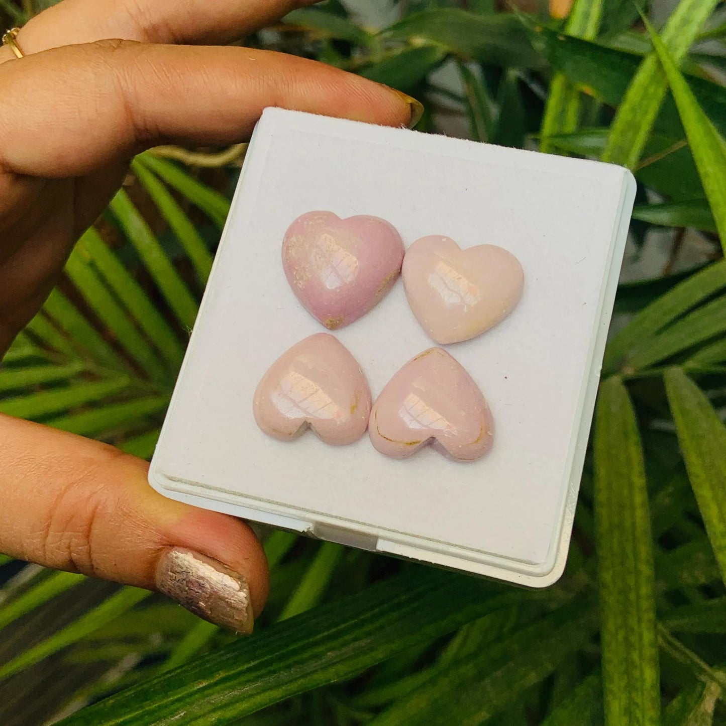 Hand holding four pink heart-shaped gemstone cabochons on a white tray, natural background