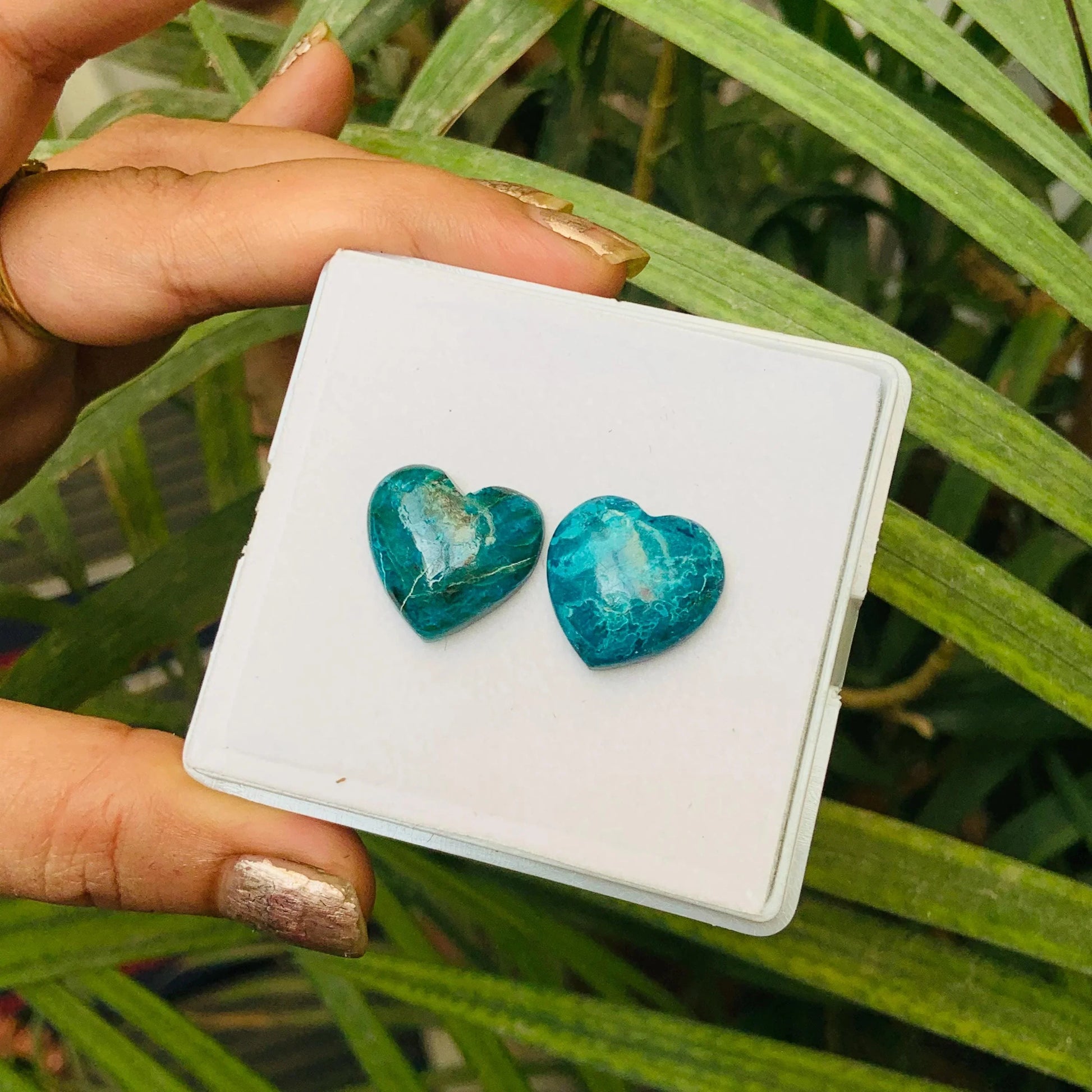 Hand holding two heart-shaped blue gemstone cabochons against green leaves