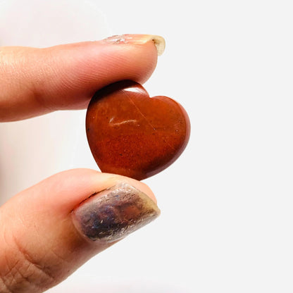 Hand holding a polished red jasper heart-shaped gemstone bead on white background