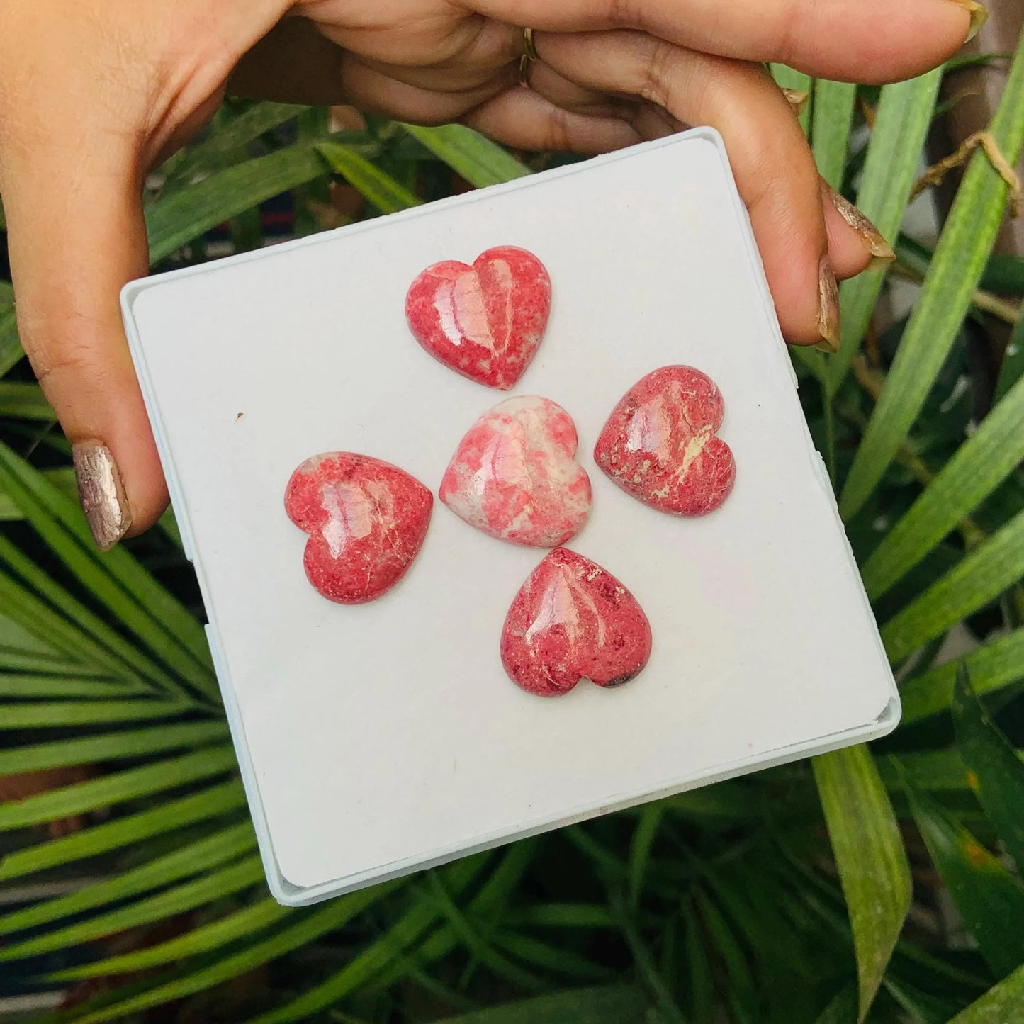 Hand holding five pink heart-shaped gemstone cabochons displayed on a white box