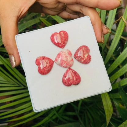 Hand holding five pink heart-shaped gemstone cabochons displayed on a white box