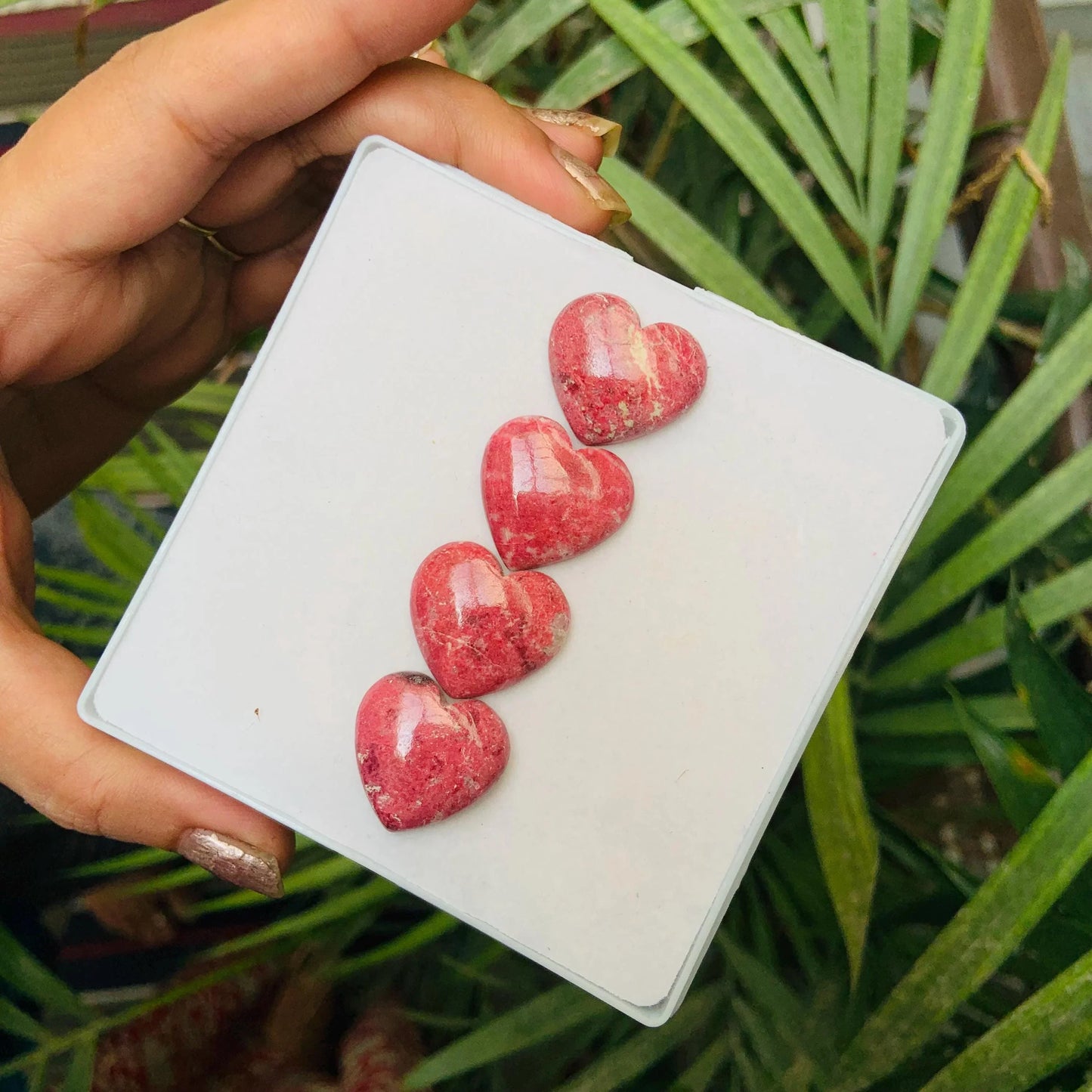 Hand holding four pink heart-shaped gemstone cabochons on a white card, greenery background