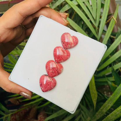 Hand holding four pink heart-shaped gemstone cabochons on a white card, greenery background